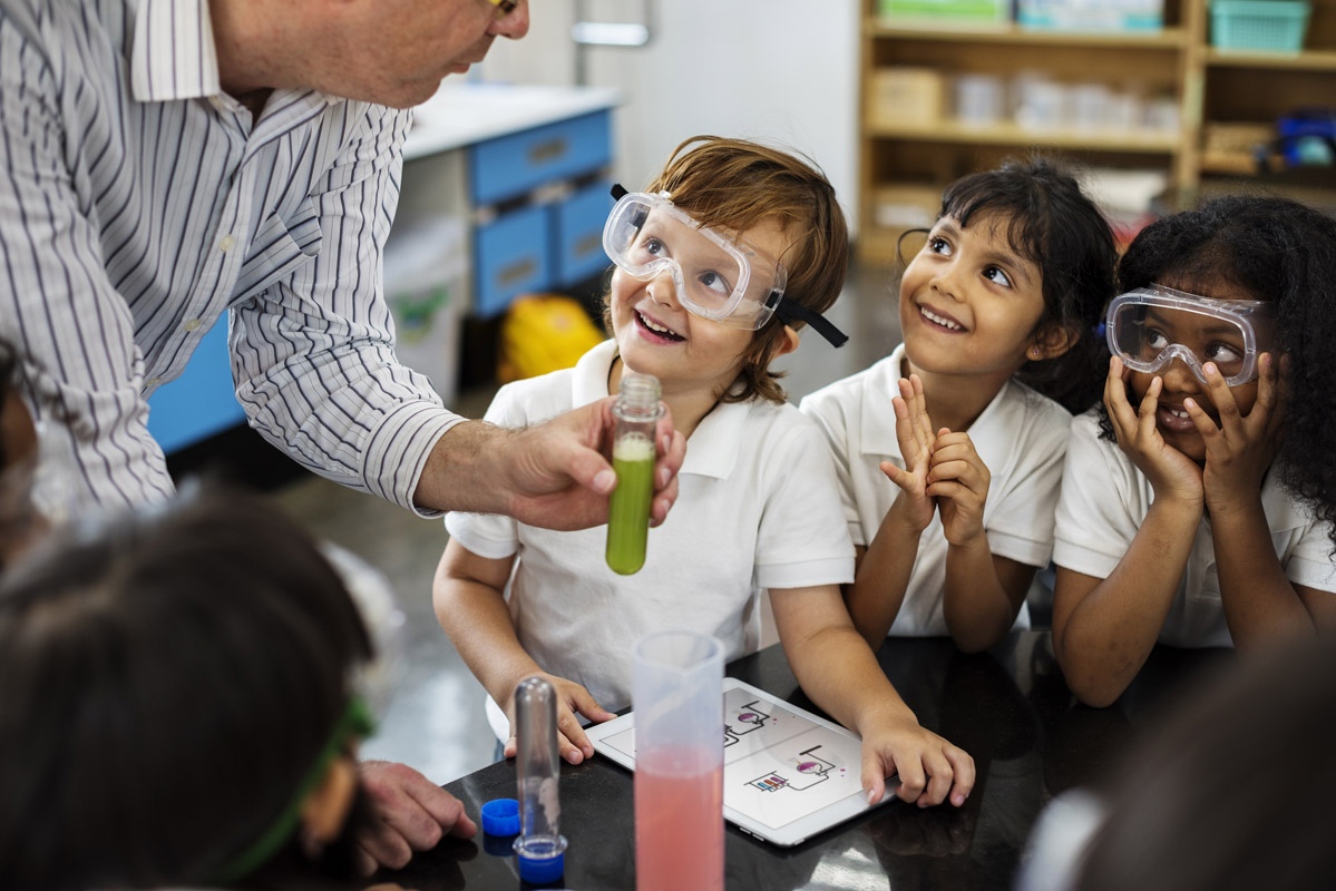 kindergarten students wearing goggles and observing a science teacher holding a test tube