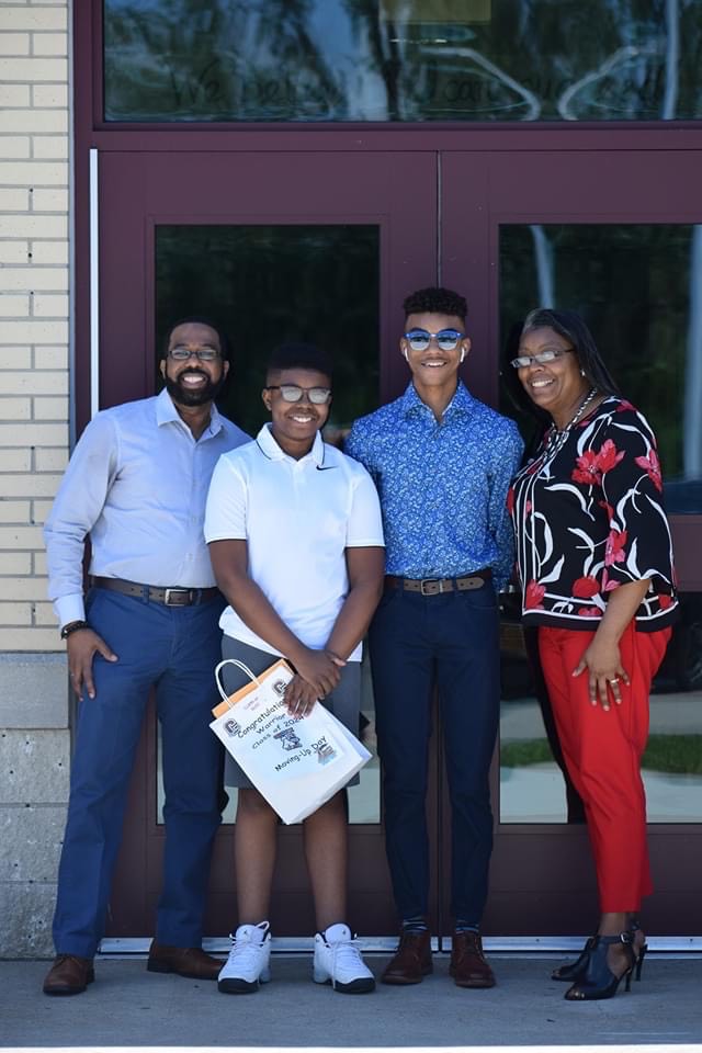 A graduating student stands with his family at the school's front entrance