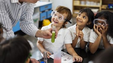 kindergarten students wearing goggles and observing a science teacher holding a test tube